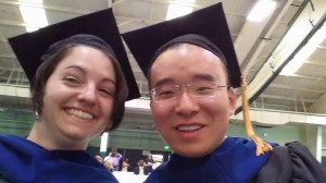 Selfie of a woman and her colleague. Both are wearing black graduation gowns with a navy blue neckpiece and black caps.