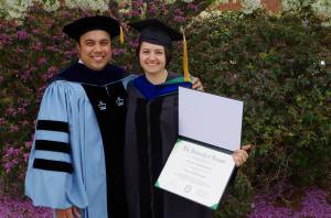 Image of a woman and her colleague taking a picture together and smiling. The woman is wearing a black graduation cap with a long yellow tassel and a black gown and is holding a diploma. Her colleague is wearing a sky blue and black gown and a black cap with a yellow tassel. In the background there are tall bushes with purple flowers.