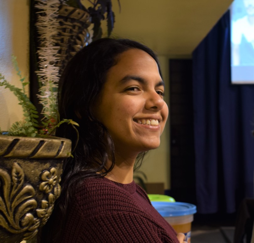 Picture of a woman smiling and leaning against a wall. The picture is from a three quarters view of her face and there are small plants on the wall next to and above her.