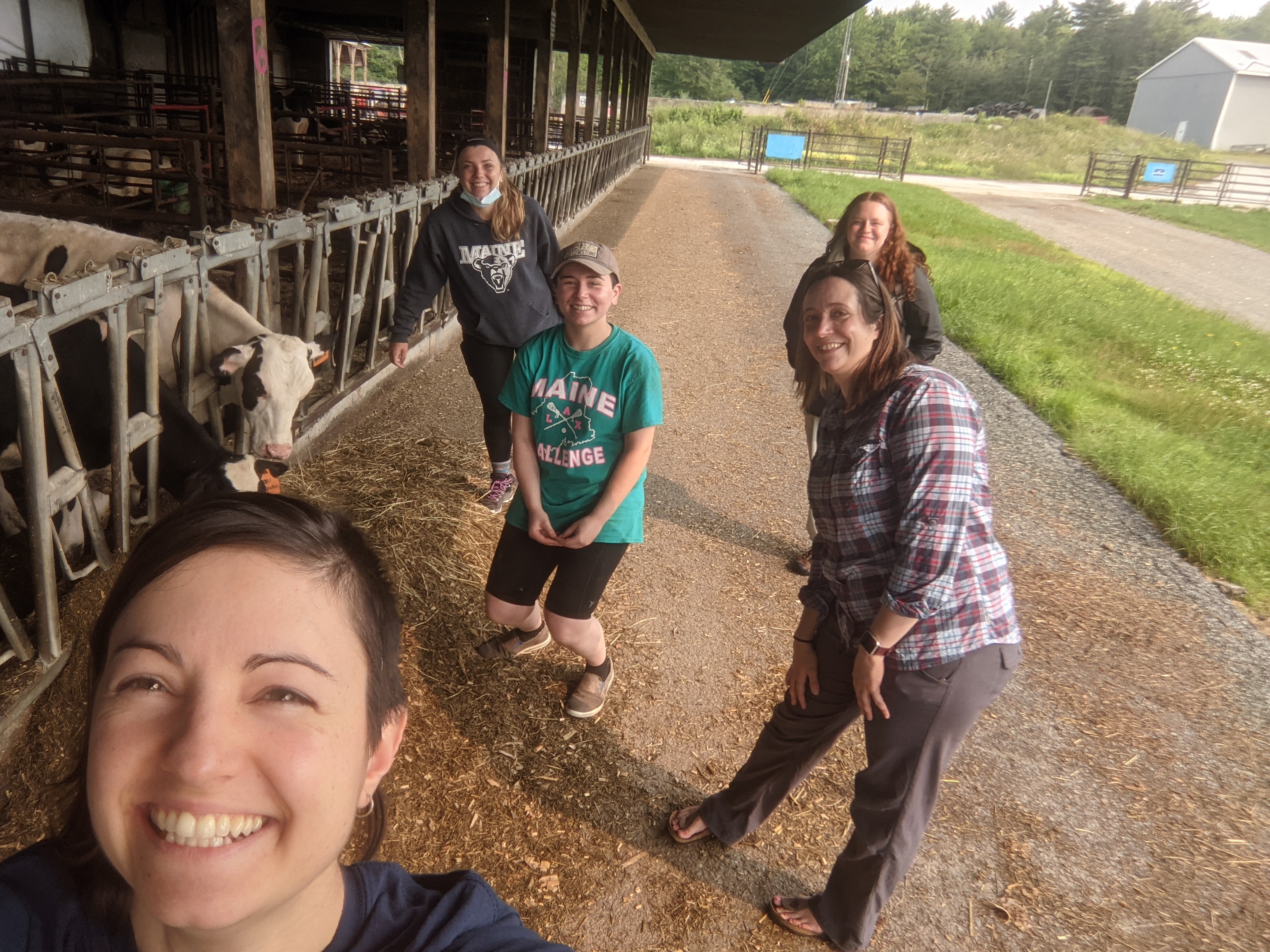 Five female researchers are standing in front of a cow feedlot posing for a photo. They are all standing a few feet apart from each other. Several cows can be seen eating next to them.