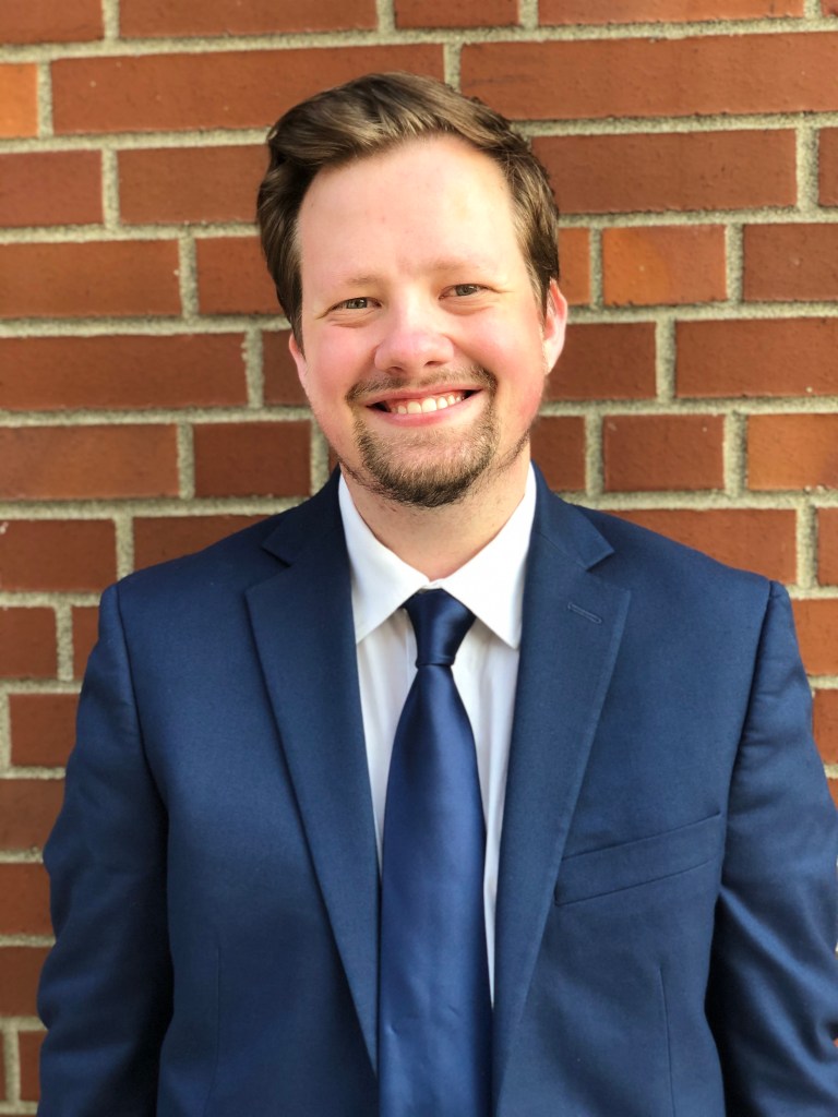Patrick is pictured here in a navy blue suit and tie against a red brick 
