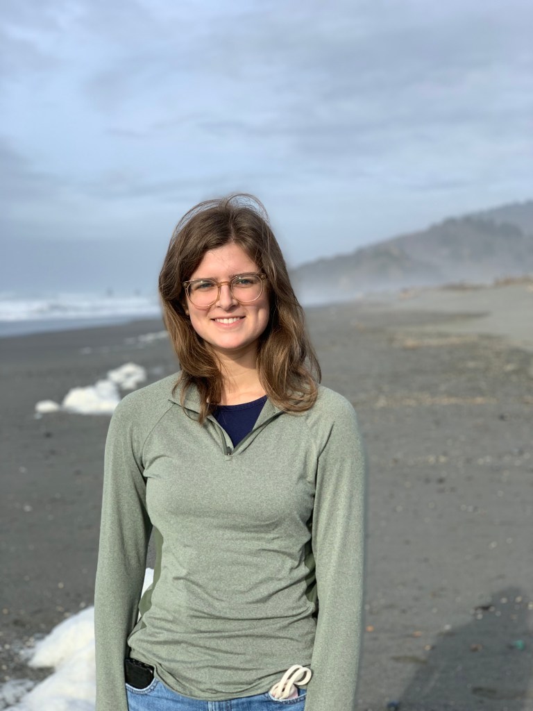 Emily Wissel on a sandy beach, with a view of the ocean and a mountainside in the background. She has glasses and is wearing a long sleeve green jacket.