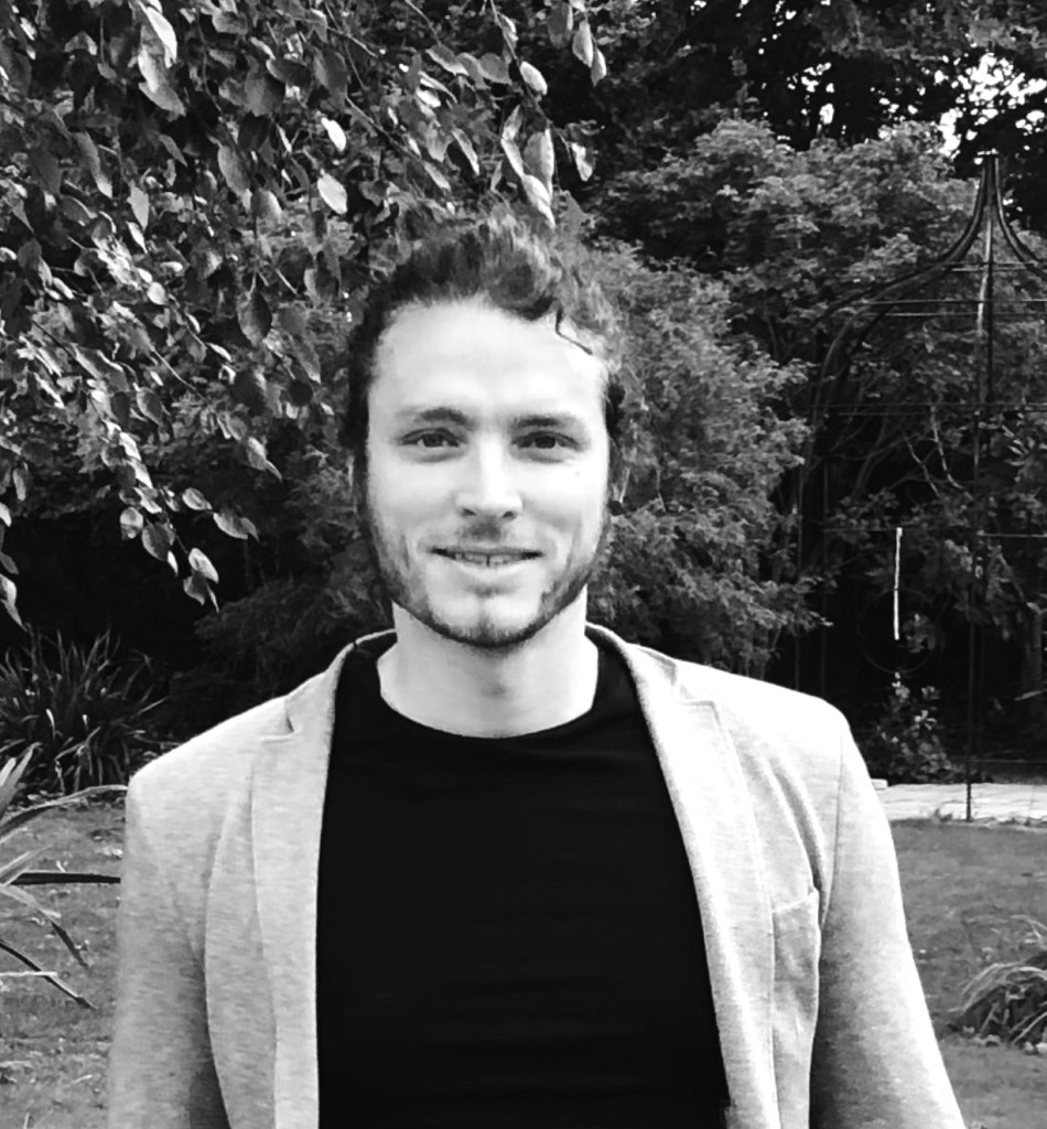 A black and white portrait of Dr. Jake Robinson, who is wearing a black shirt and light sportscoat over it. Jake is outside in front of some bushes.