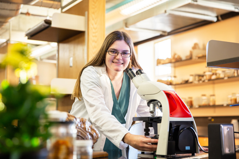 Sarah Hosler posing at a microscope, wearing a lab coat, standing in a research lab