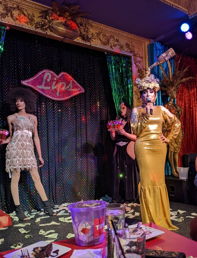 Several drag queens in elaborate gowns, hairstyles, and makeup, standing on a stage.