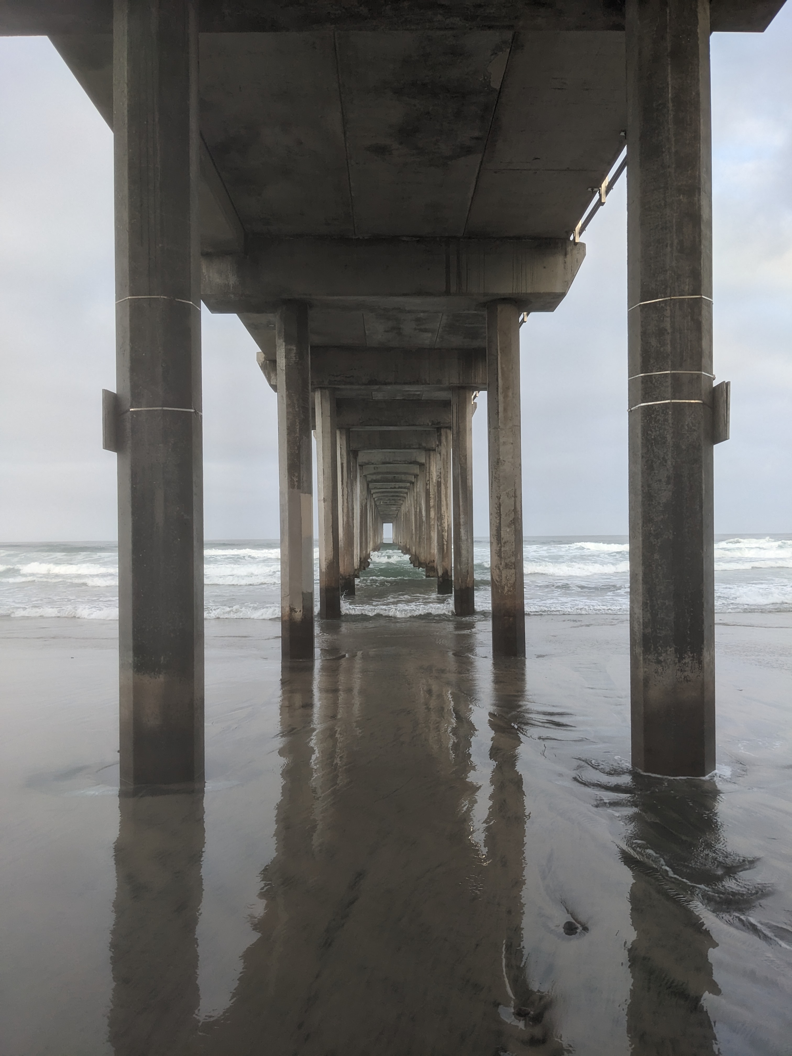 A view down the underside of a concrete pier, with breaking waves and cloudy sky in the background and wet sand in the foreground.