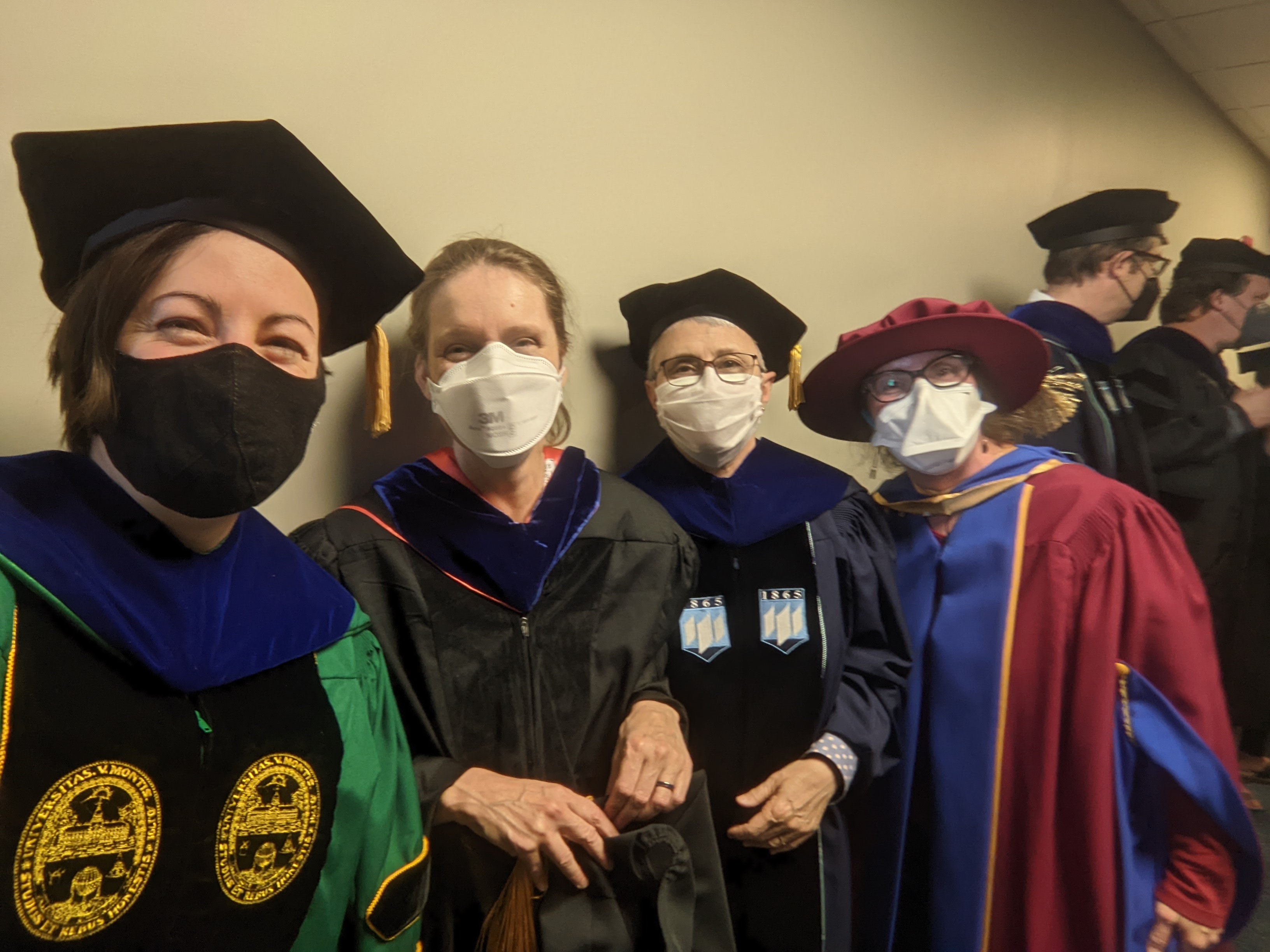 Four professors wearing full regalia, as well as face masks, posing for a photo in a hallway.