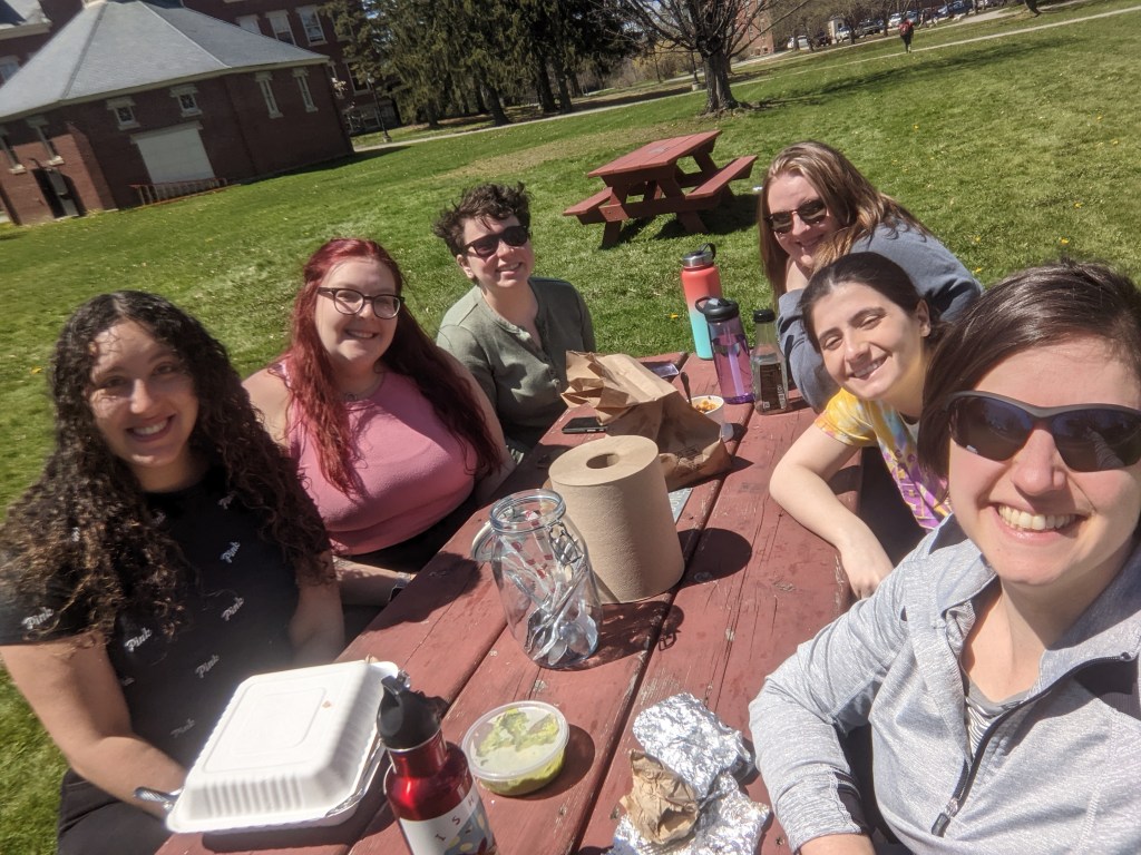 Six women sitting at a picnic table eating lunch outside, and smiling at the camera.