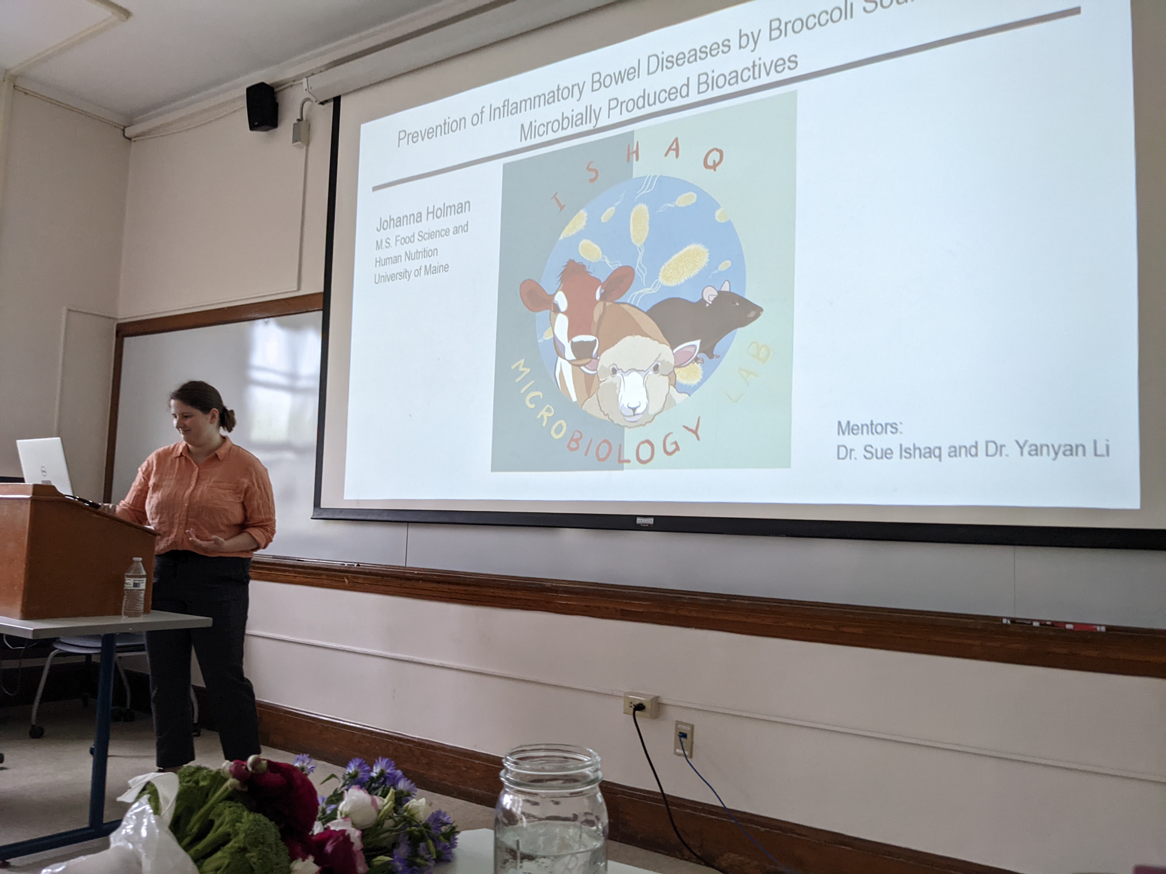 Johanna Holman standing at a wooden podium with a white board and a projection screen behind her. Johanna is presenting her master's thesis, and the title slide of the presentation is showing on the screen.