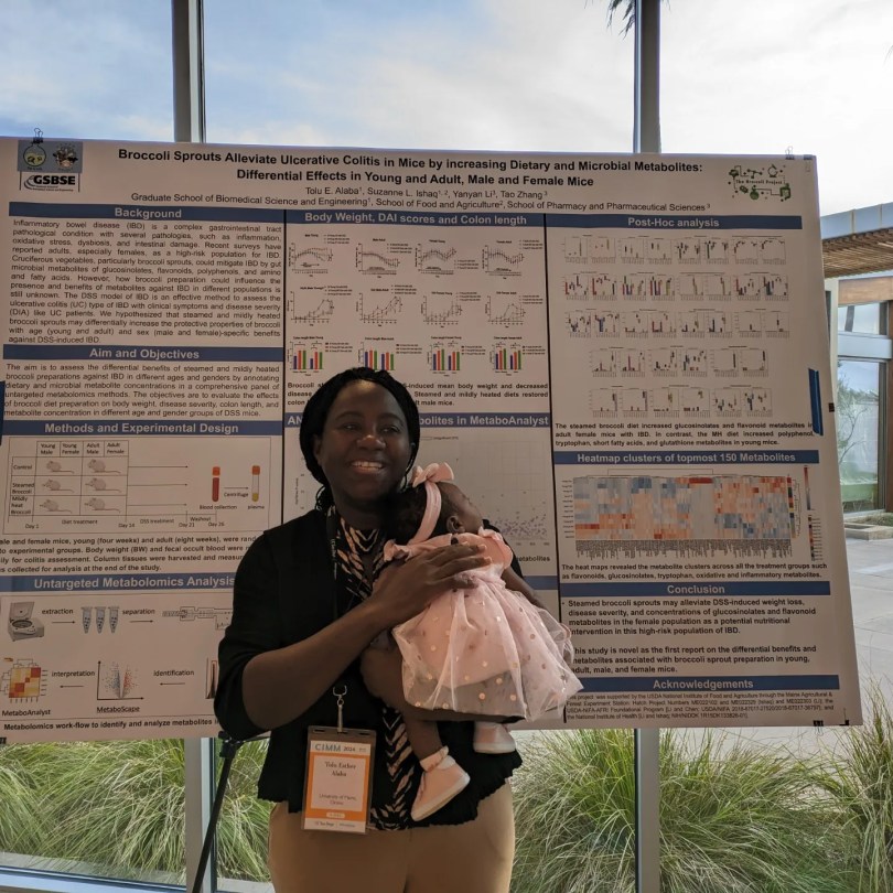 Tolu Esther Alaba is standing in front of a science conference poster, smiling, and holding her infant.