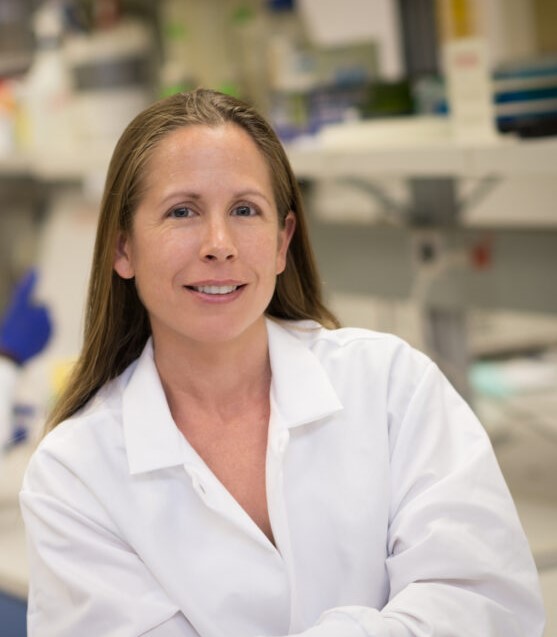 Dr. Kelly Baker posing for a professional headshot. She's wearing a white lab coat and she's standing in a microbiology lab which is blurred out in the background.