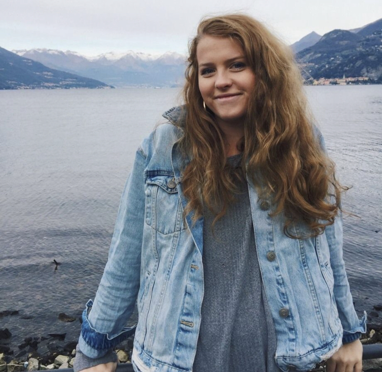 Mary Coughter is wearing a grey shirt and denim jacket, and is standing on a rocky shoreline in front of a large body of water with mountains in the background.