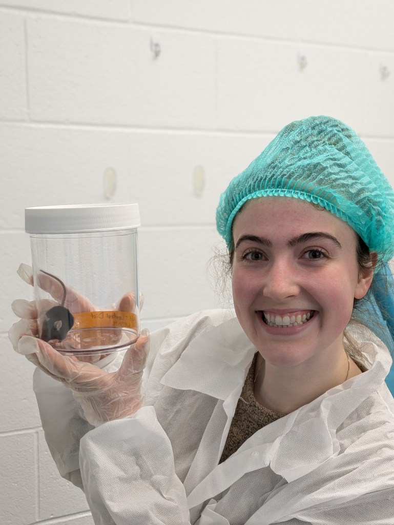 Ashley is holding up a plastic jar with a research mouse in it.