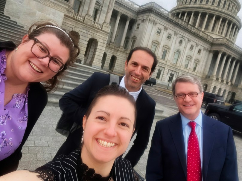 Four people in suits posing for a selfie in front of the US Capitol Building.